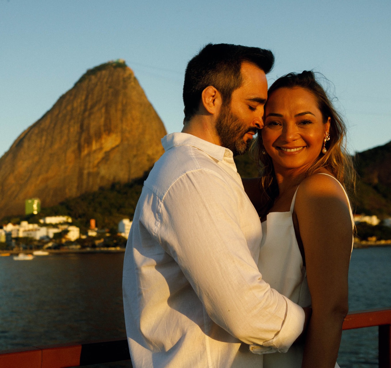 Casal posa junto à proa da escuna ao pôr do sol, com o Pão de Açúcar iluminado ao fundo na Baía de Guanabara